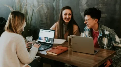 Easy online side hustles for college students you can start today. Image shows three college-aged women sharing a table in a coworking space, while working with their laptops.