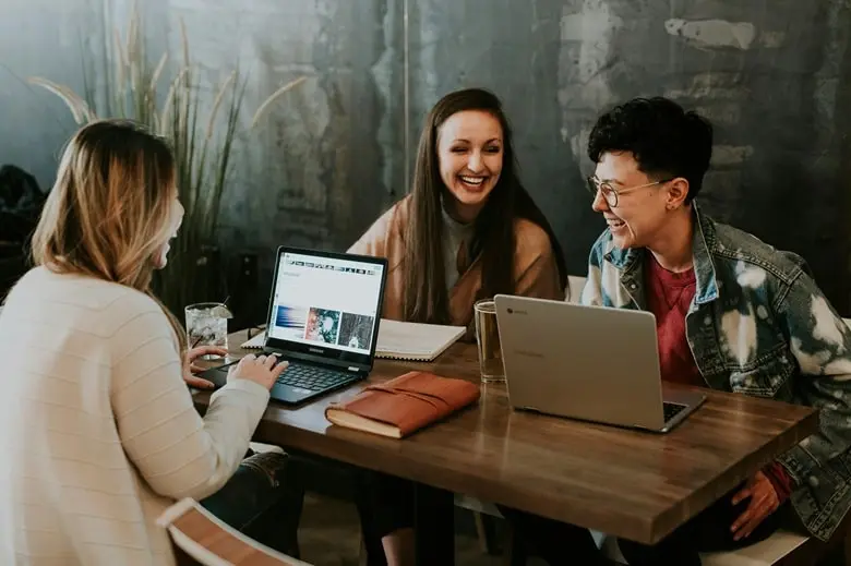Easy online side hustles for college students you can start today. Image shows three college-aged women sharing a table in a coworking space, while working with their laptops.